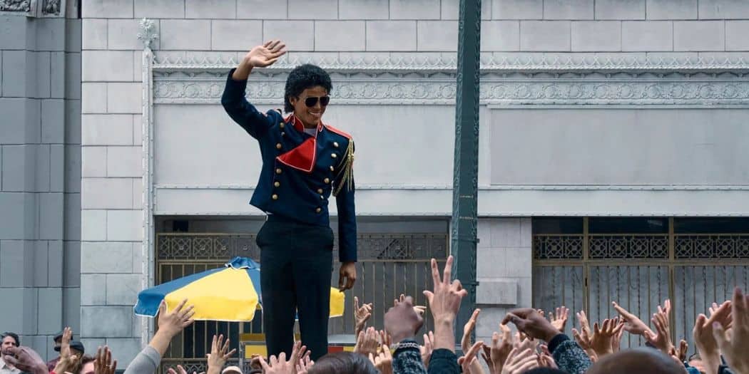 Jaafar Jackson en veste militaire bleu et rouge, lunettes de soleil, saluant du haut d'une estrade une foule nombreuse dont les mains se tendent vers lui.