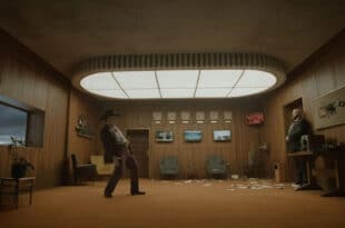 Dans Digger, Tom Cruise porte un chapeau noir et un costume bordeaux dans un bureau boisé, face à un homme près de la porte.