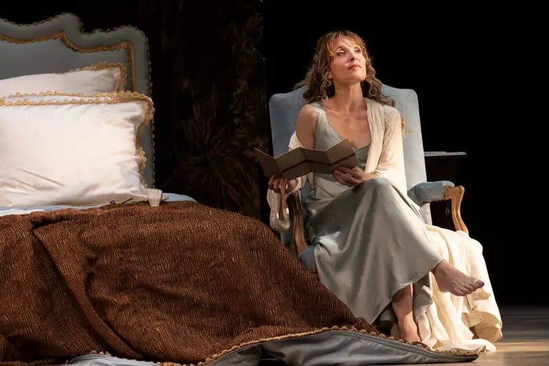 Alix Poisson en robe claire, assise près d’un lit, un livre ouvert dans les mains.